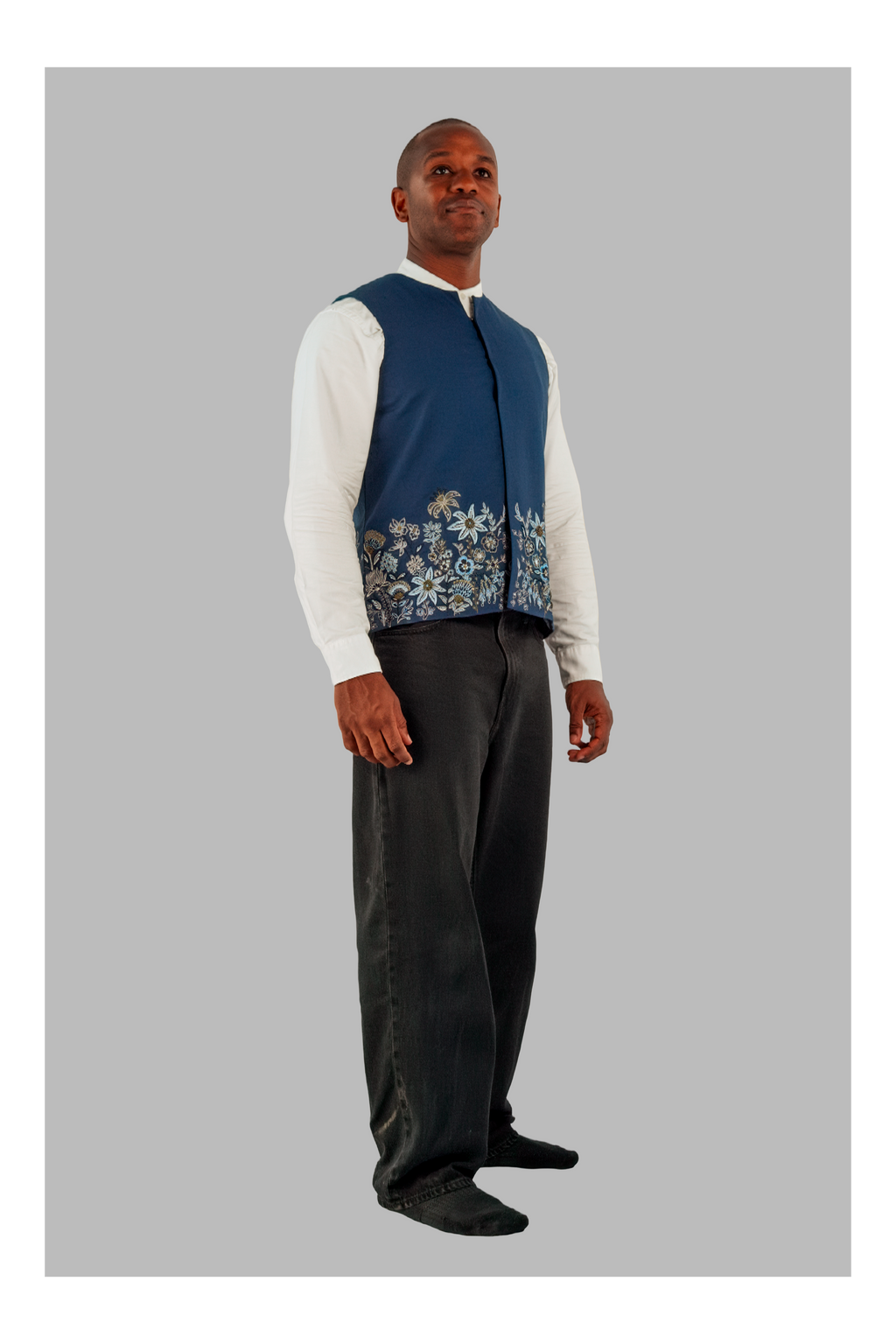 Man wearing a blue vest with floral pattern over a white shirt and dark pants on a gray background