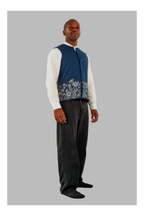 Man wearing a blue vest with floral pattern over a white shirt and dark pants on a gray background