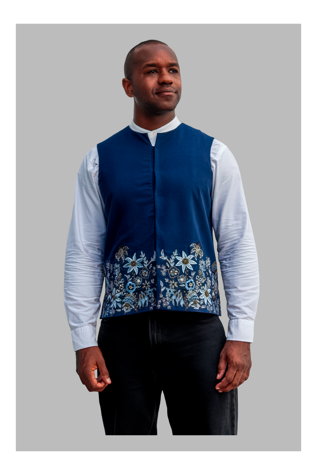 Man wearing a blue embroidered vest over a white shirt with black pants on a gray background