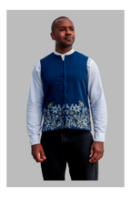 Man wearing a blue embroidered vest over a white shirt with black pants on a gray background