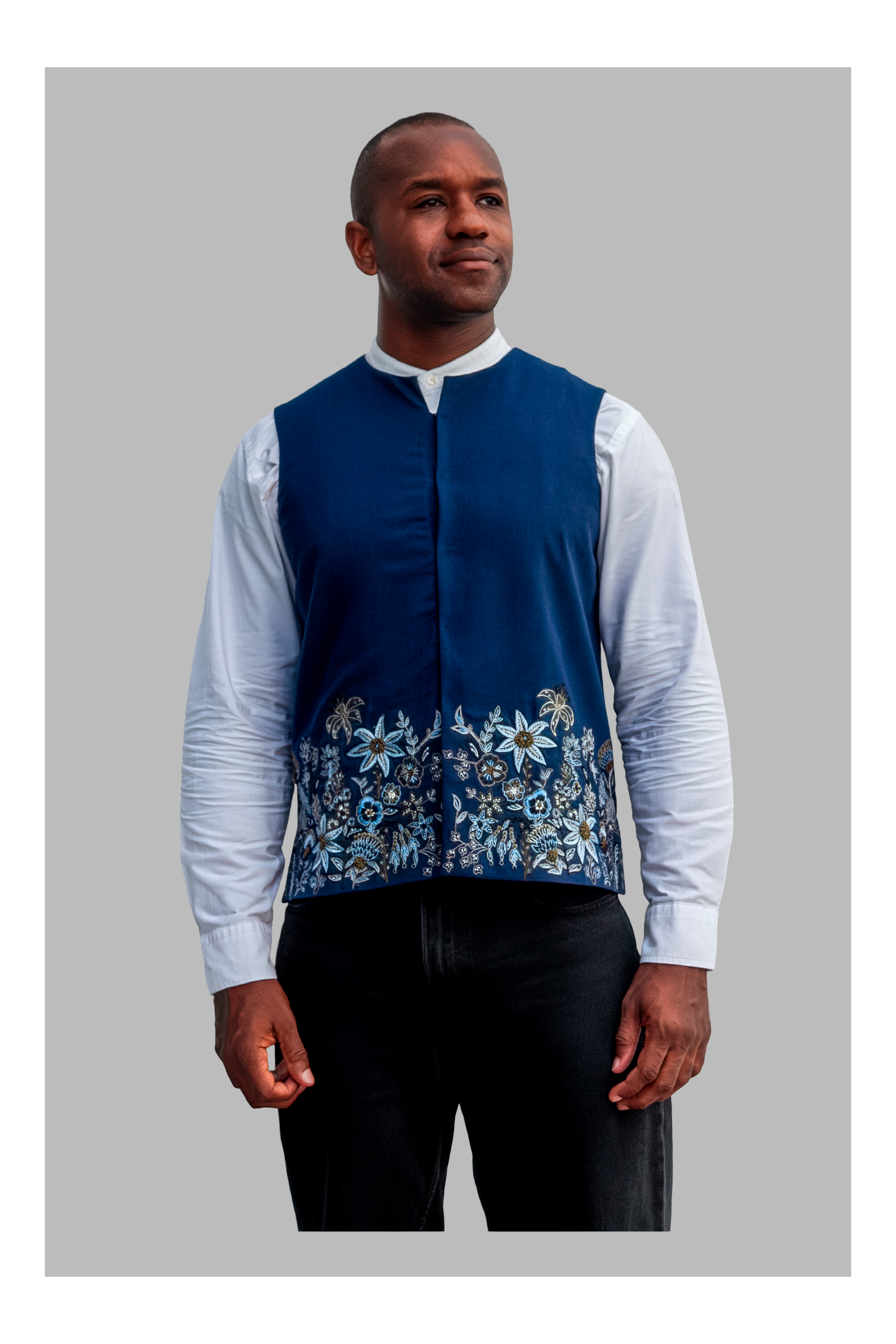 Man wearing a blue embroidered vest over a white shirt with black pants on a gray background