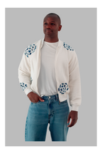 Man wearing a white bomber jacket with blue patterns, white t-shirt, and blue jeans on a gray background