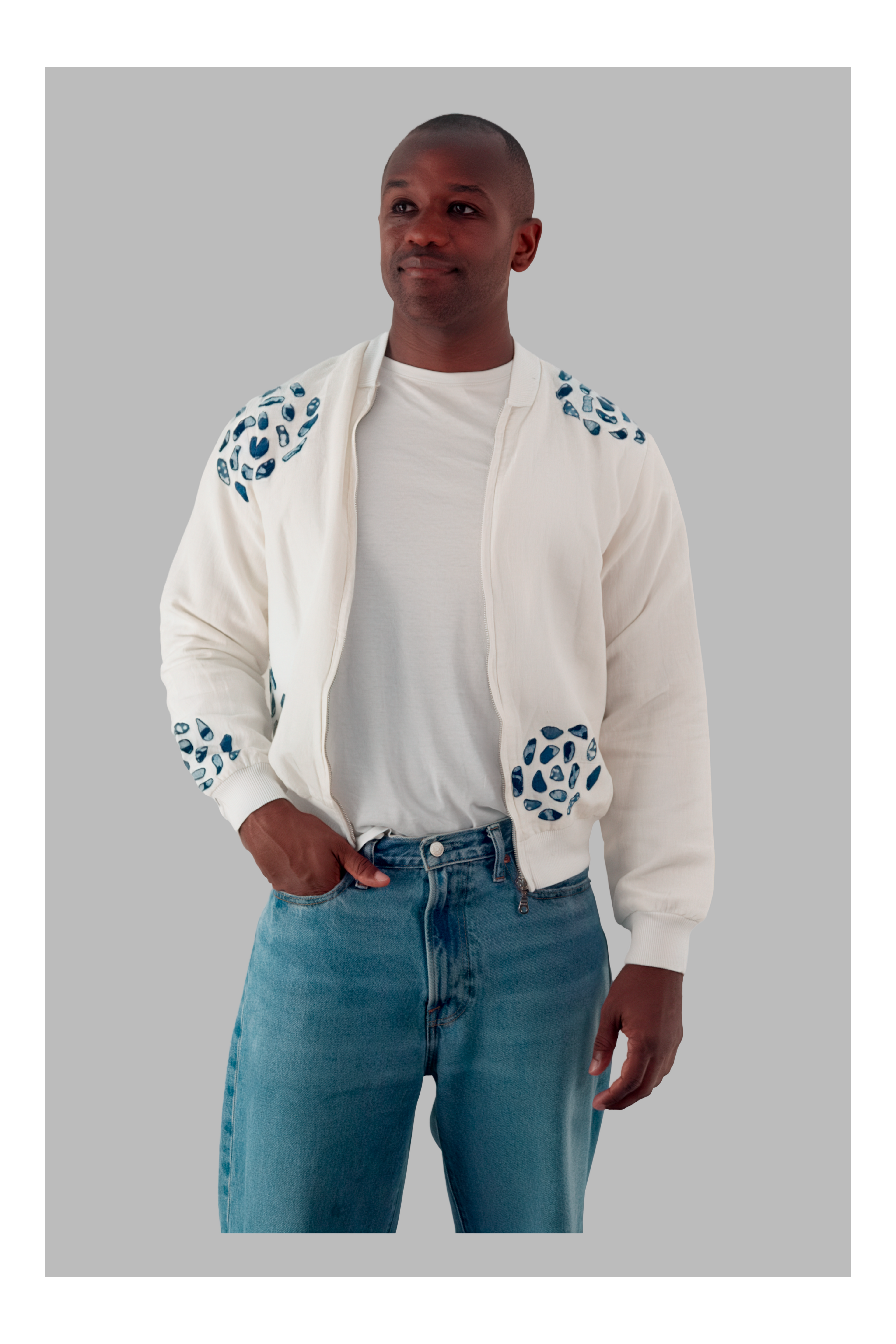 Man wearing a white bomber jacket with blue patterns, white t-shirt, and blue jeans on a gray background