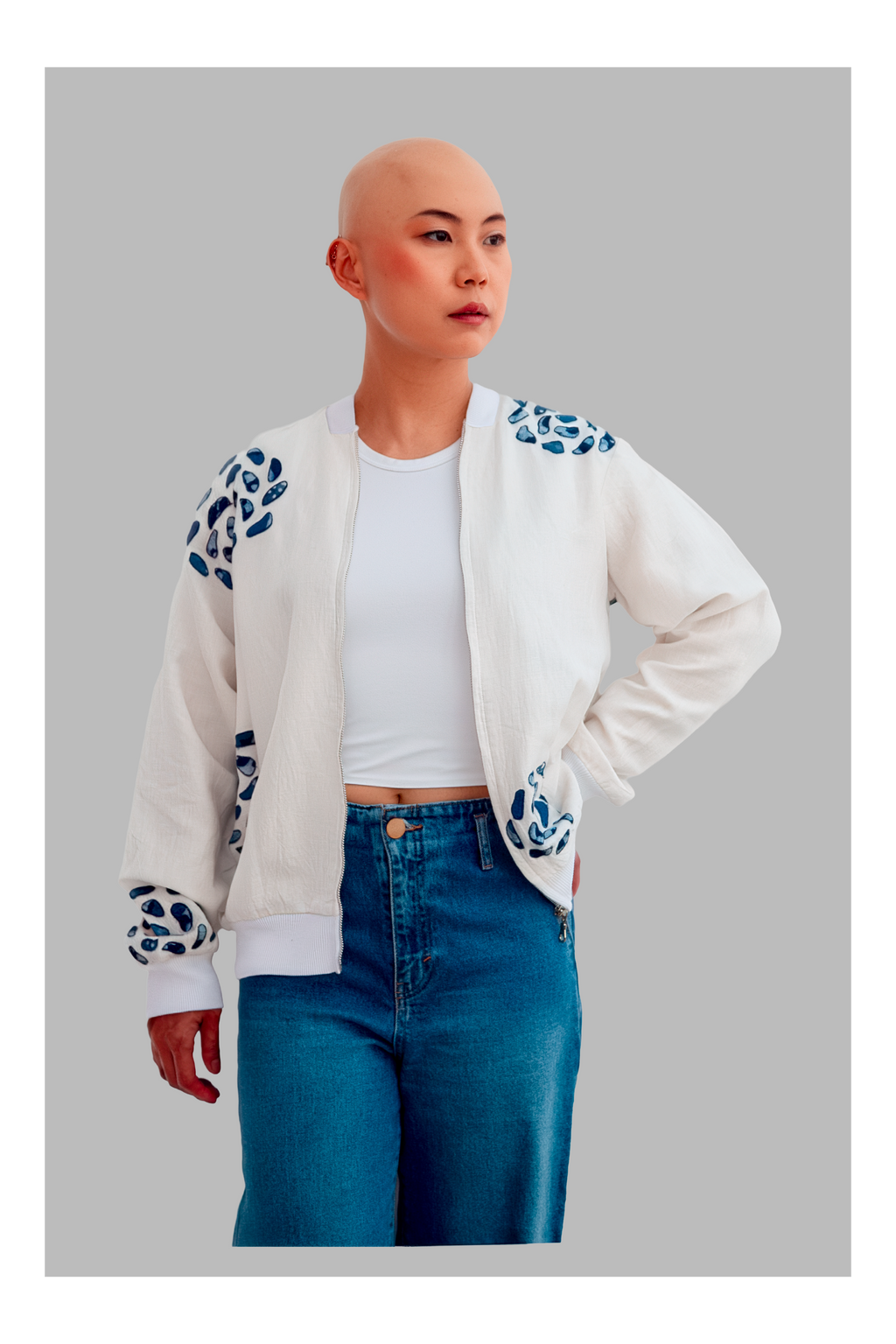 Female wearing a white bomber jacket with blue patterns, white shirt, and blue jeans on a gray background