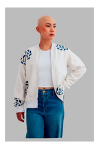 Female wearing a white bomber jacket with blue patterns, white shirt, and blue jeans on a gray background