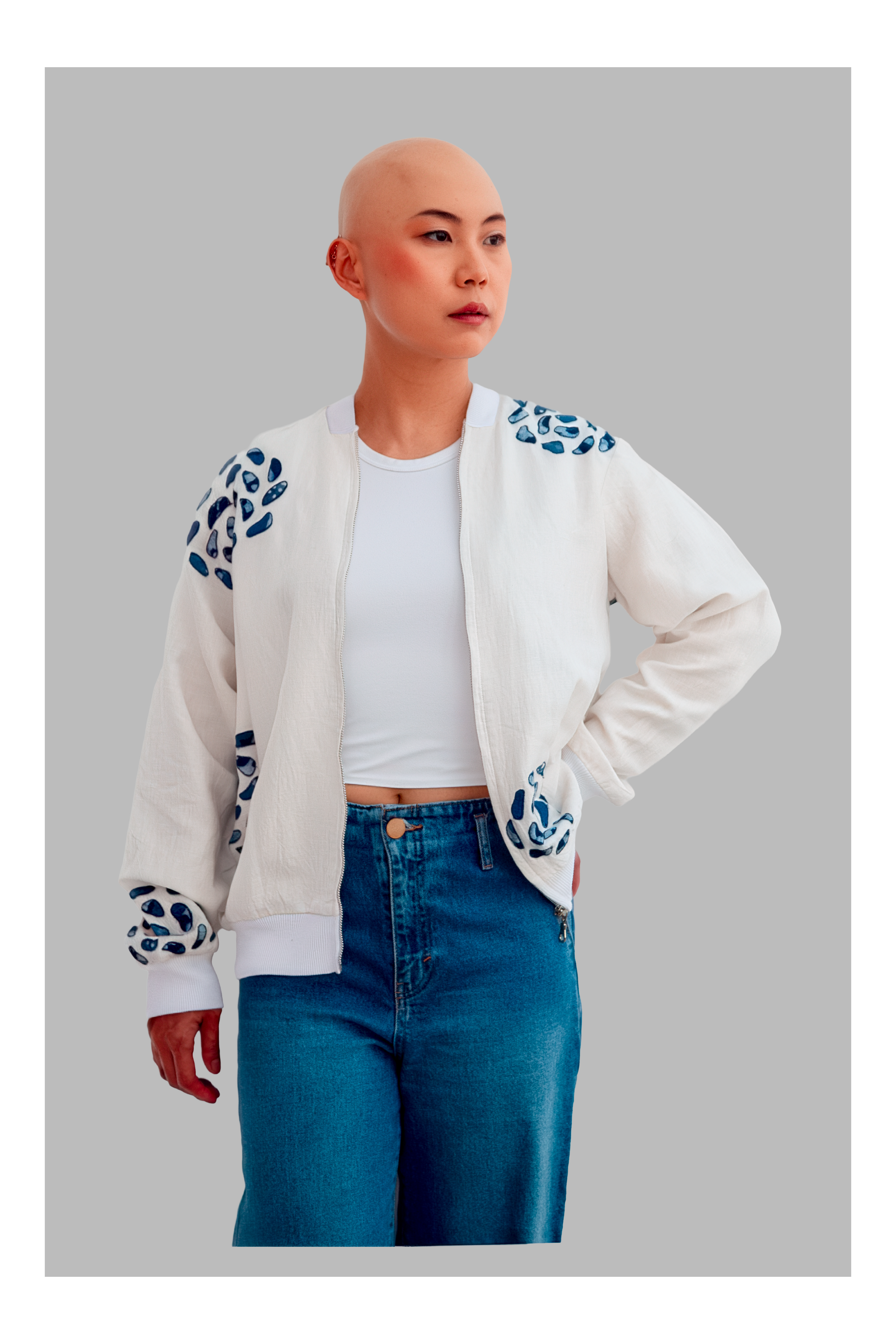 Female wearing a white bomber jacket with blue patterns, white shirt, and blue jeans on a gray background